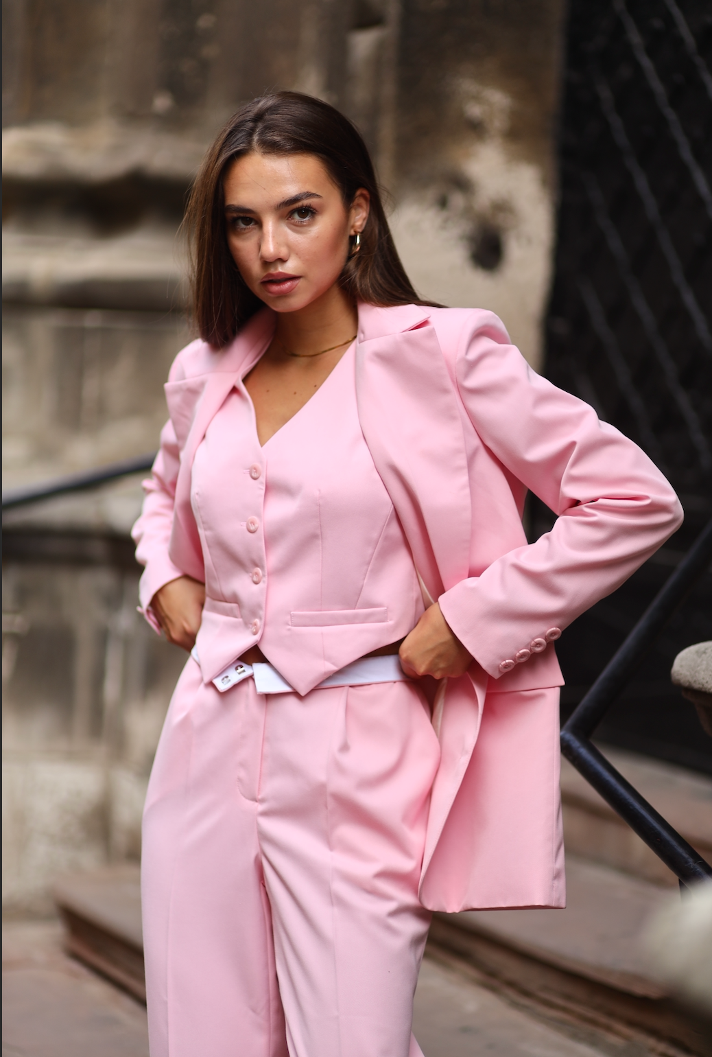 A woman wearing pink rose colored straight trousers with a matching waistcoat and skirt, standing on a sunset-lit street.