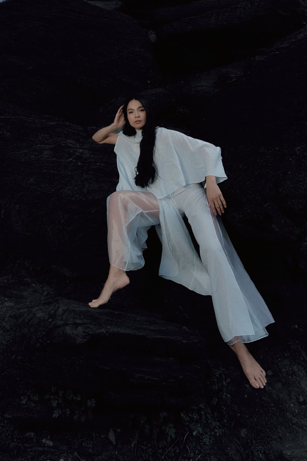Woman wearing a white silk blouse and trousers with transparent details sitting on a dark textured surface