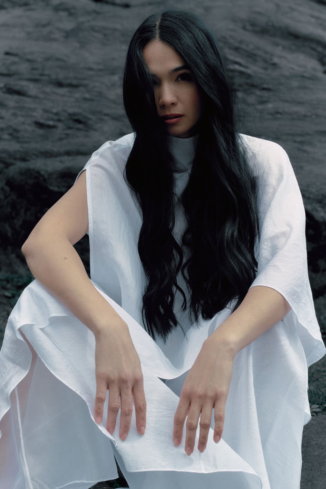 Woman wearing a white silk blouse and trousers with transparent details sitting on a dark textured surface