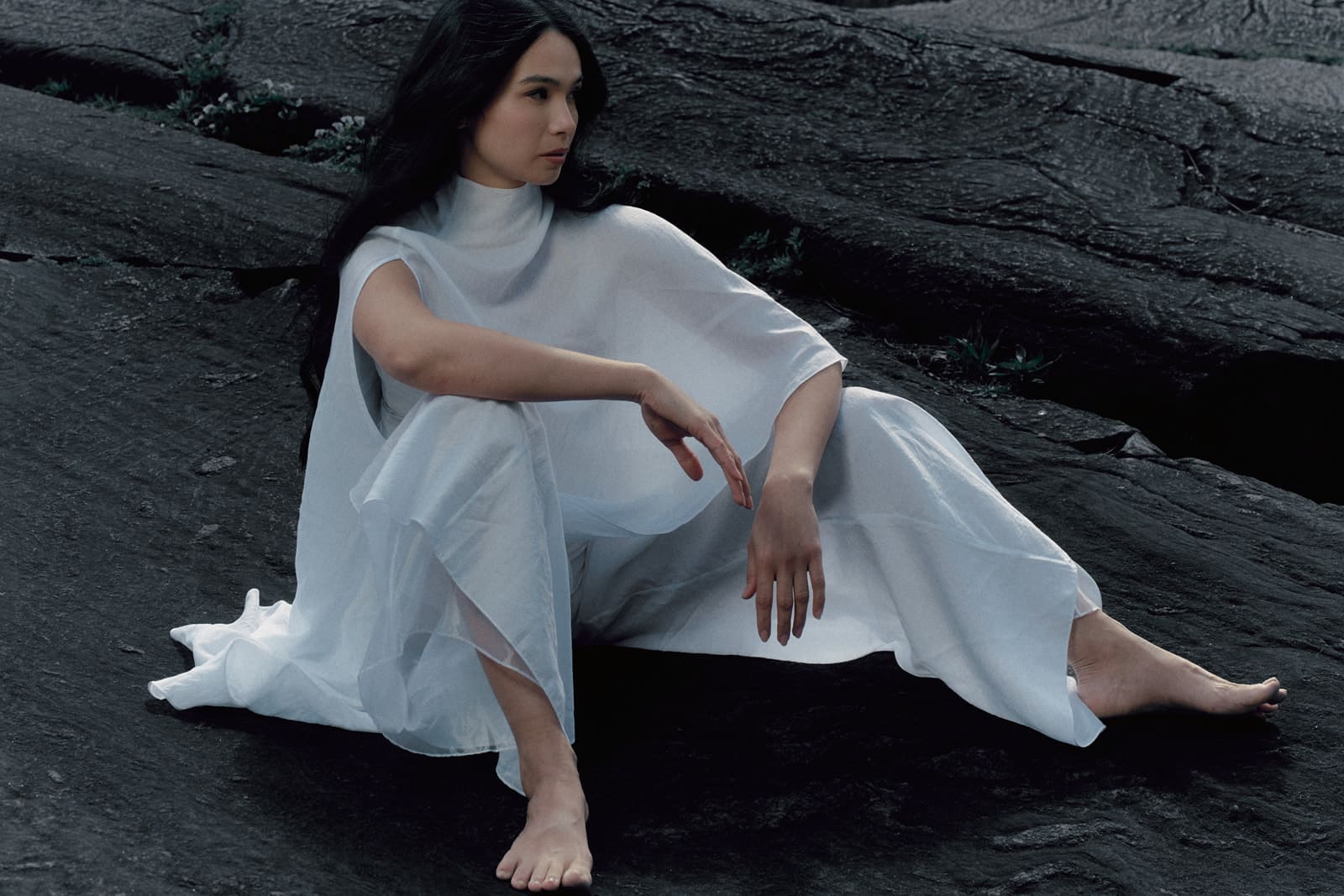 Woman wearing a white silk blouse and trousers with transparent details sitting on a dark textured surface