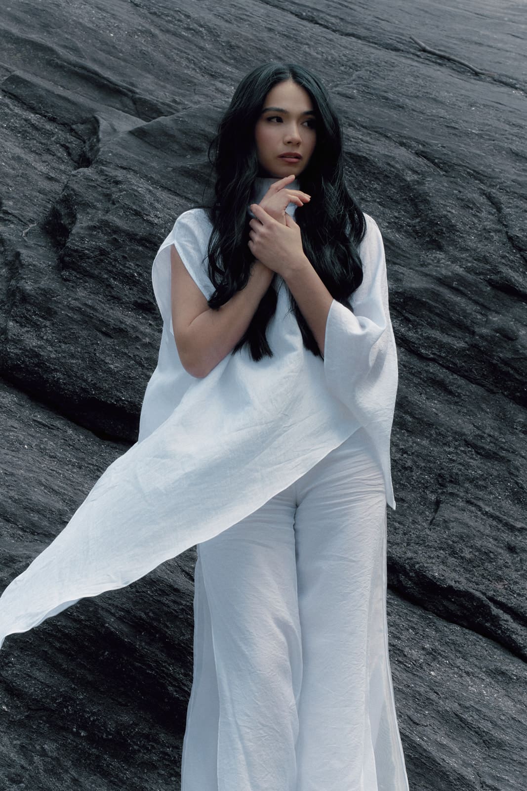 Woman wearing a white silk blouse and trousers with transparent details sitting on a dark textured surface