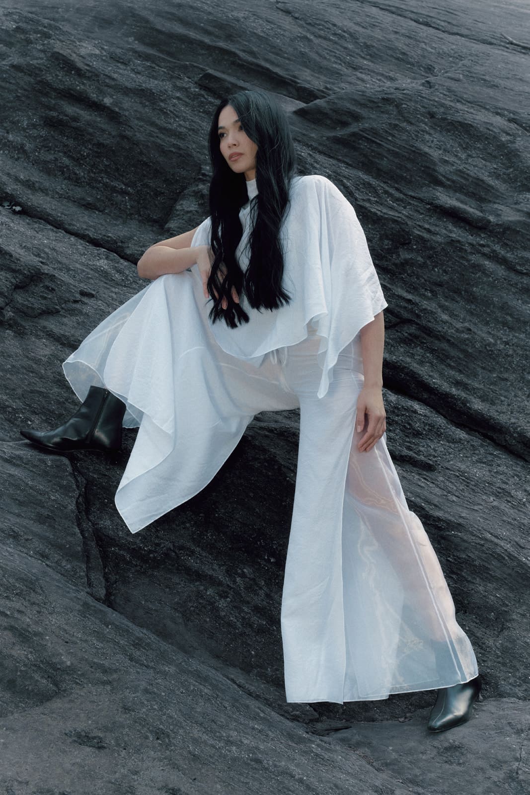 Woman wearing a white silk blouse and trousers with transparent details sitting on a dark textured surface