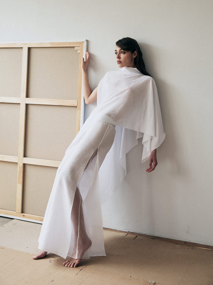 Woman in a white outfit standing next to a large wooden frame against a plain wall.