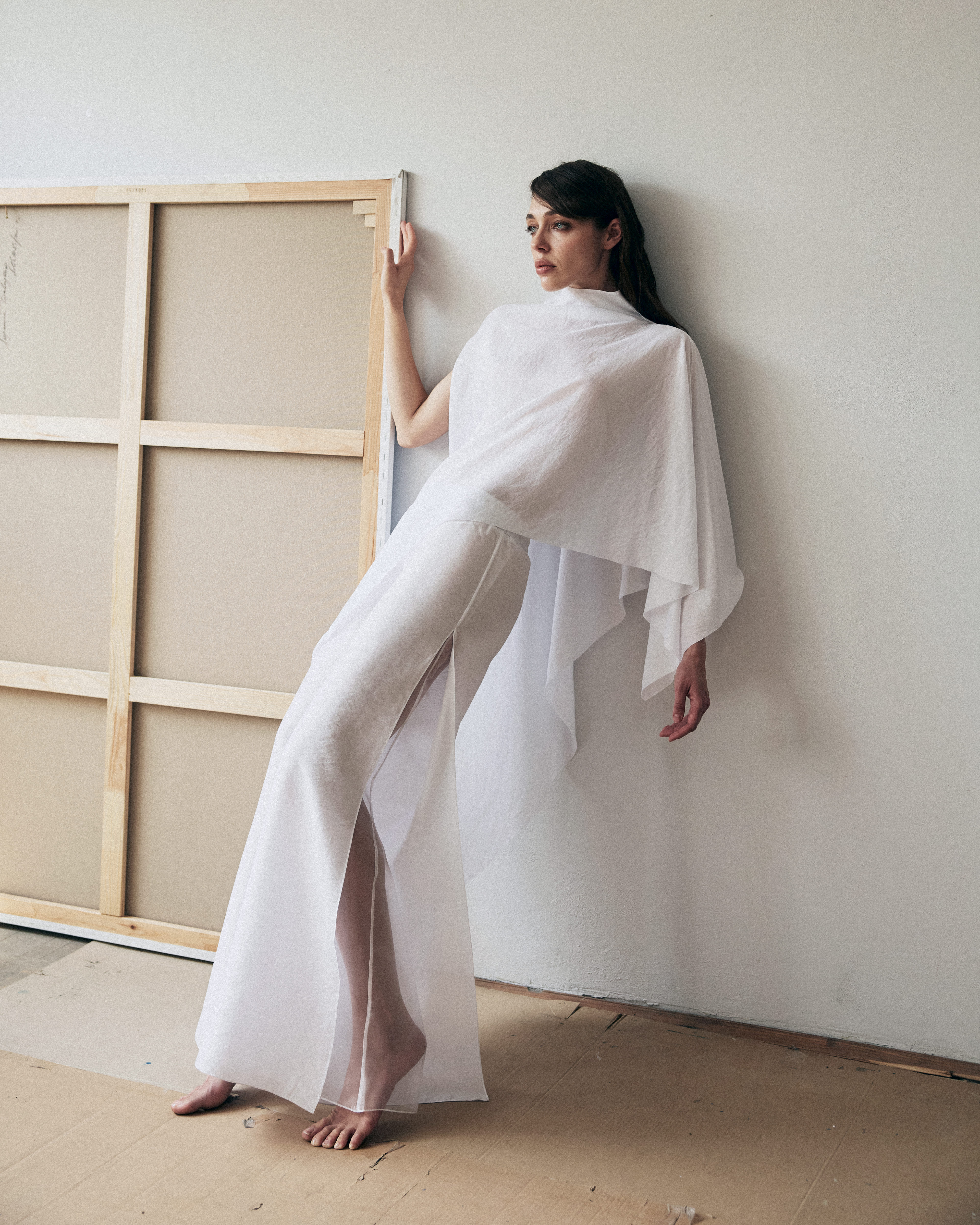 Woman in a white outfit standing next to a large wooden frame against a plain wall.