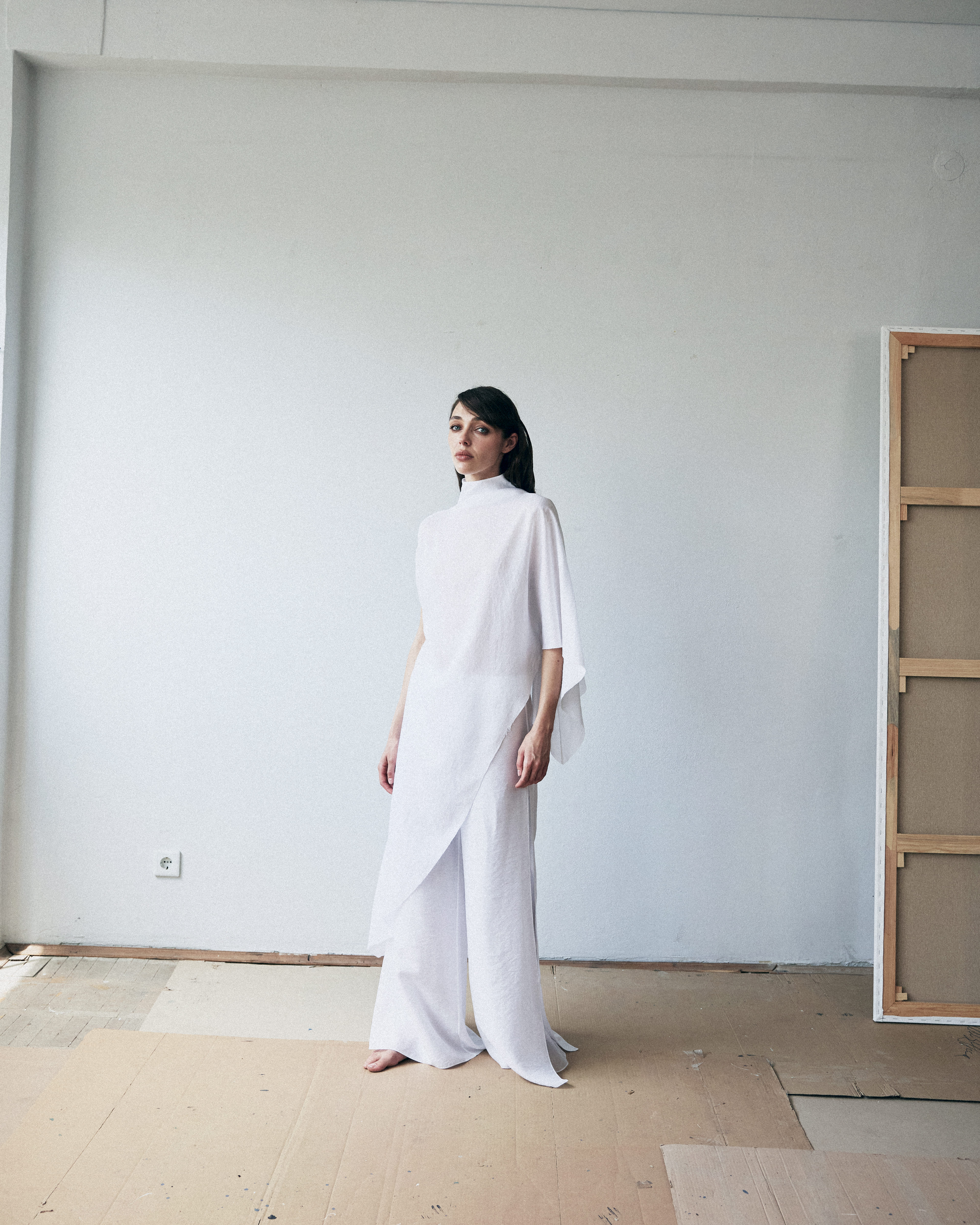 Woman in a white outfit standing in a minimalistic room with a plain wall and wooden floor.