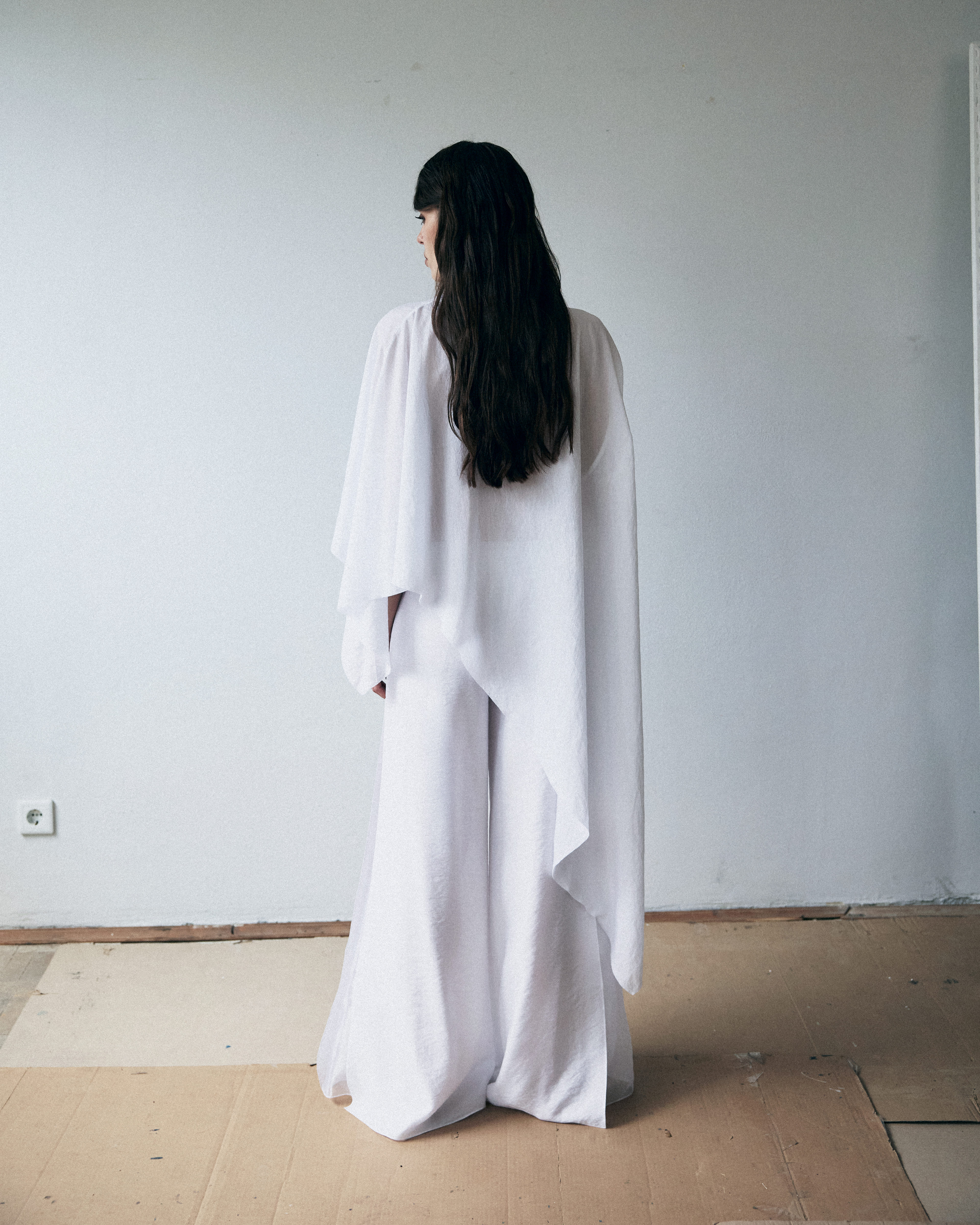 Person wearing a long white dress standing against a plain wall.