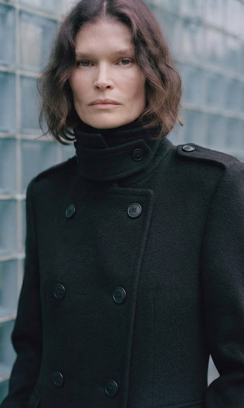 A woman wearing a dark charcoal black double-breasted coat with a high folded collar, standing in front of a glass block wall.