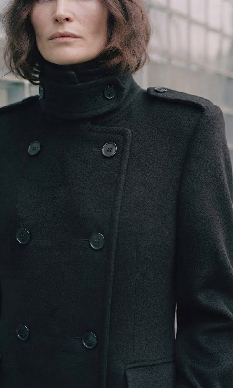 A close-up of a model wearing a dark charcoal gray double-breasted coat with a high folded collar and dark buttons.