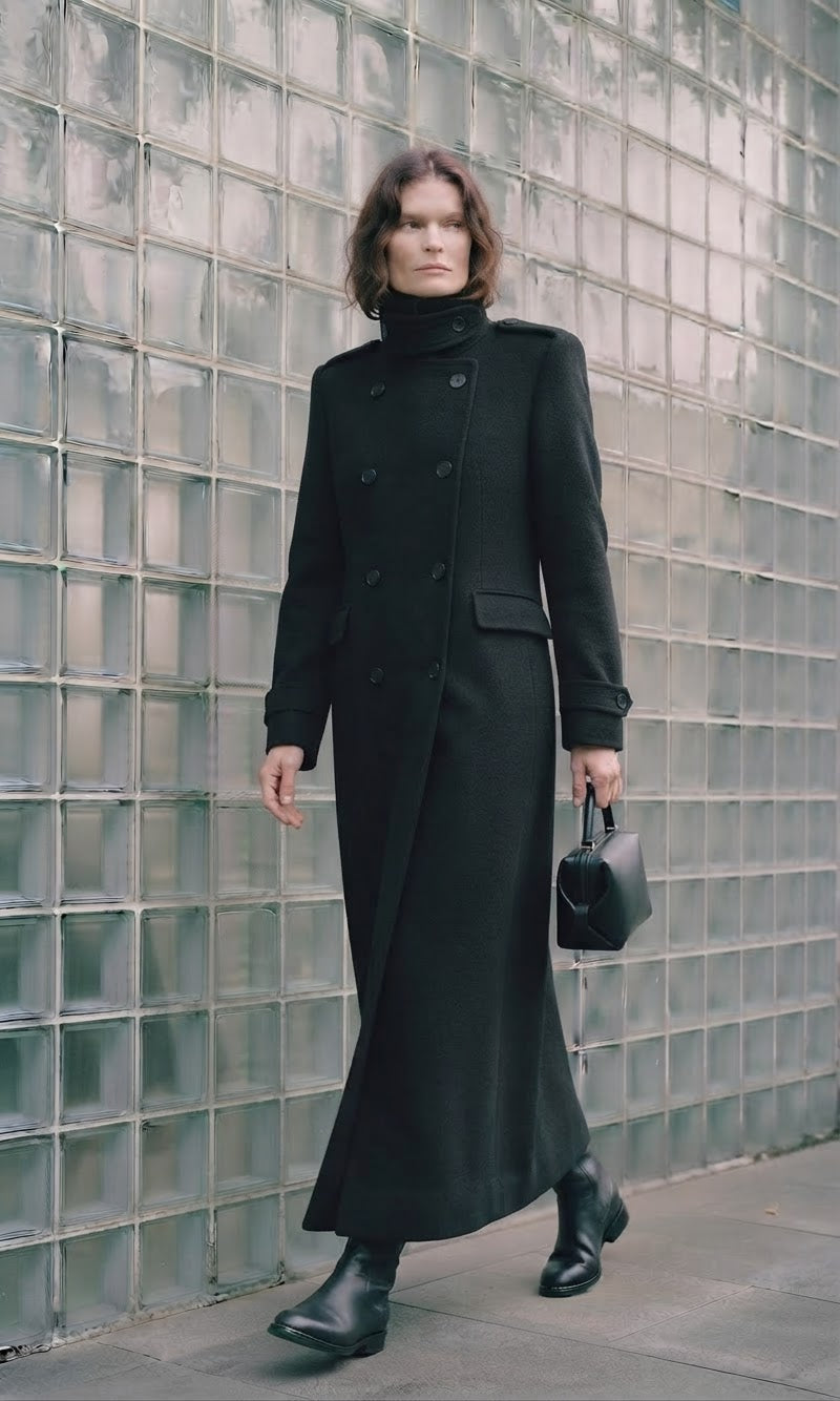 A woman wearing a long black double-breasted coat with a high mock neck, holding a small black handbag, standing in front of a glass block wall.