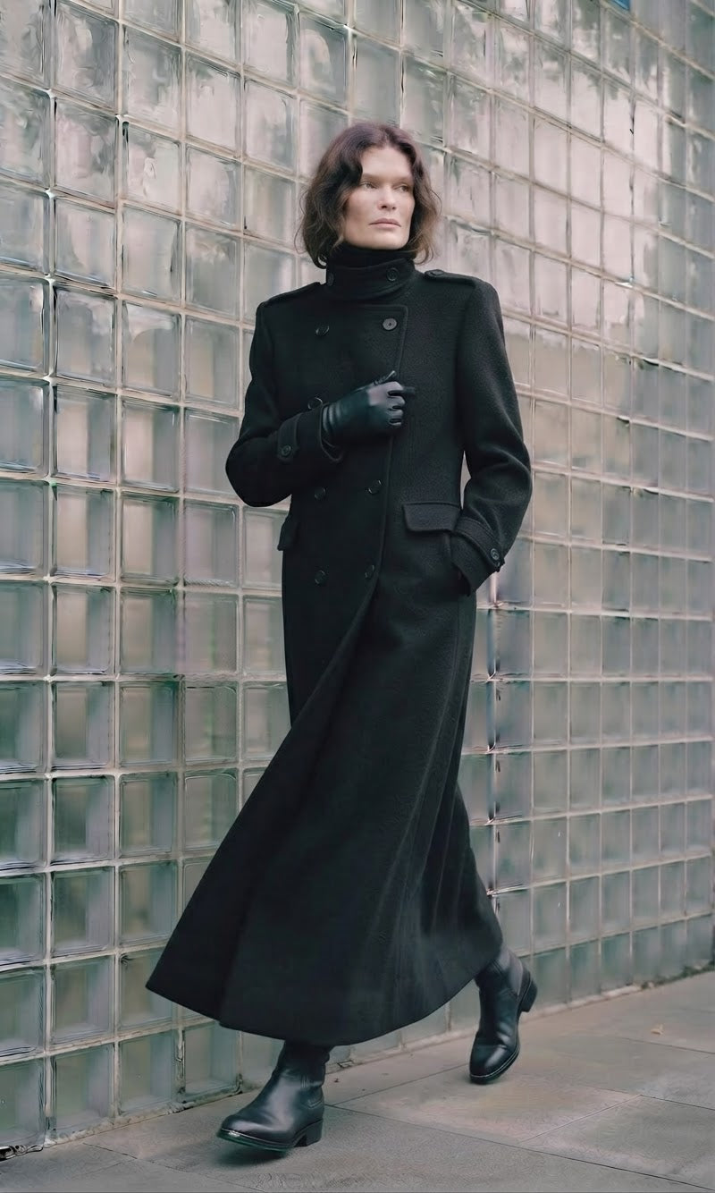 A model wearing a long black double-breasted coat, black gloves, and black ankle boots, posing in front of a glass block wall.